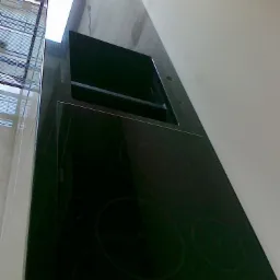 Czarny, błyszczący blat z wbudowaną płytą indukcyjną i zlewem, widok z dołu pod kątem, fragment okna w tle.