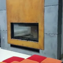 Nowoczesny kominek z obudową z betonu architektonicznego i rdzawego metalu, z prostokątnym wkładem kominkowym i widocznymi kratkami wentylacyjnymi, otoczony kolorowymi pufami.