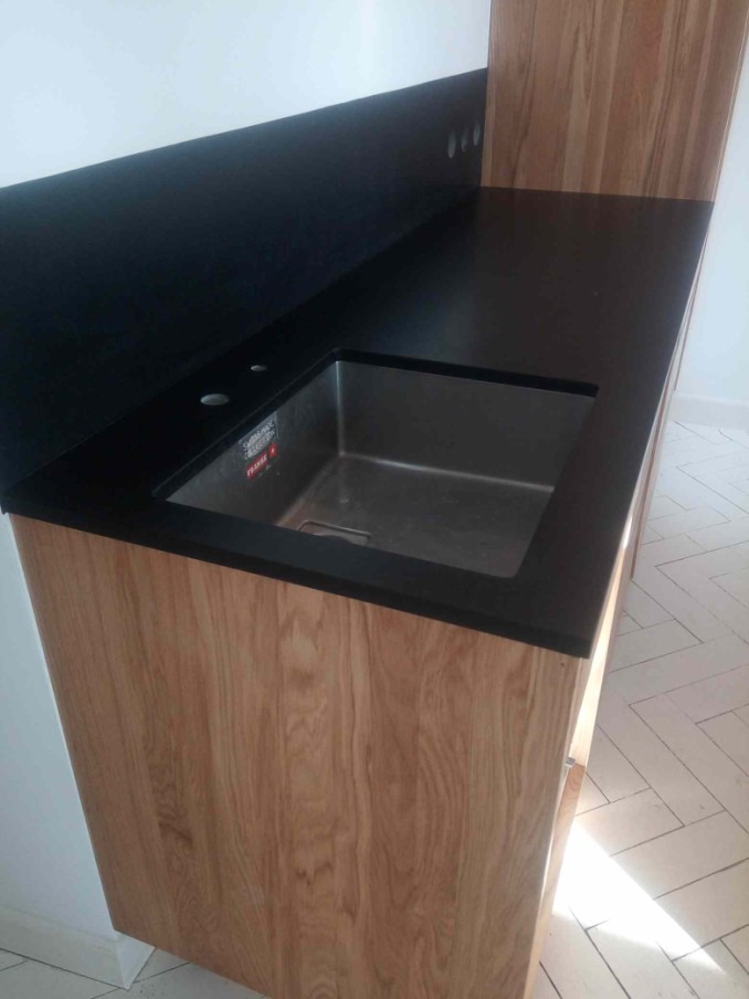 Czarny, matowy blat z wyciętym otworem na zlew, zamontowany na szafce z okleiną imitującą jasne drewno, widoczne otwory na baterię i dozownik.