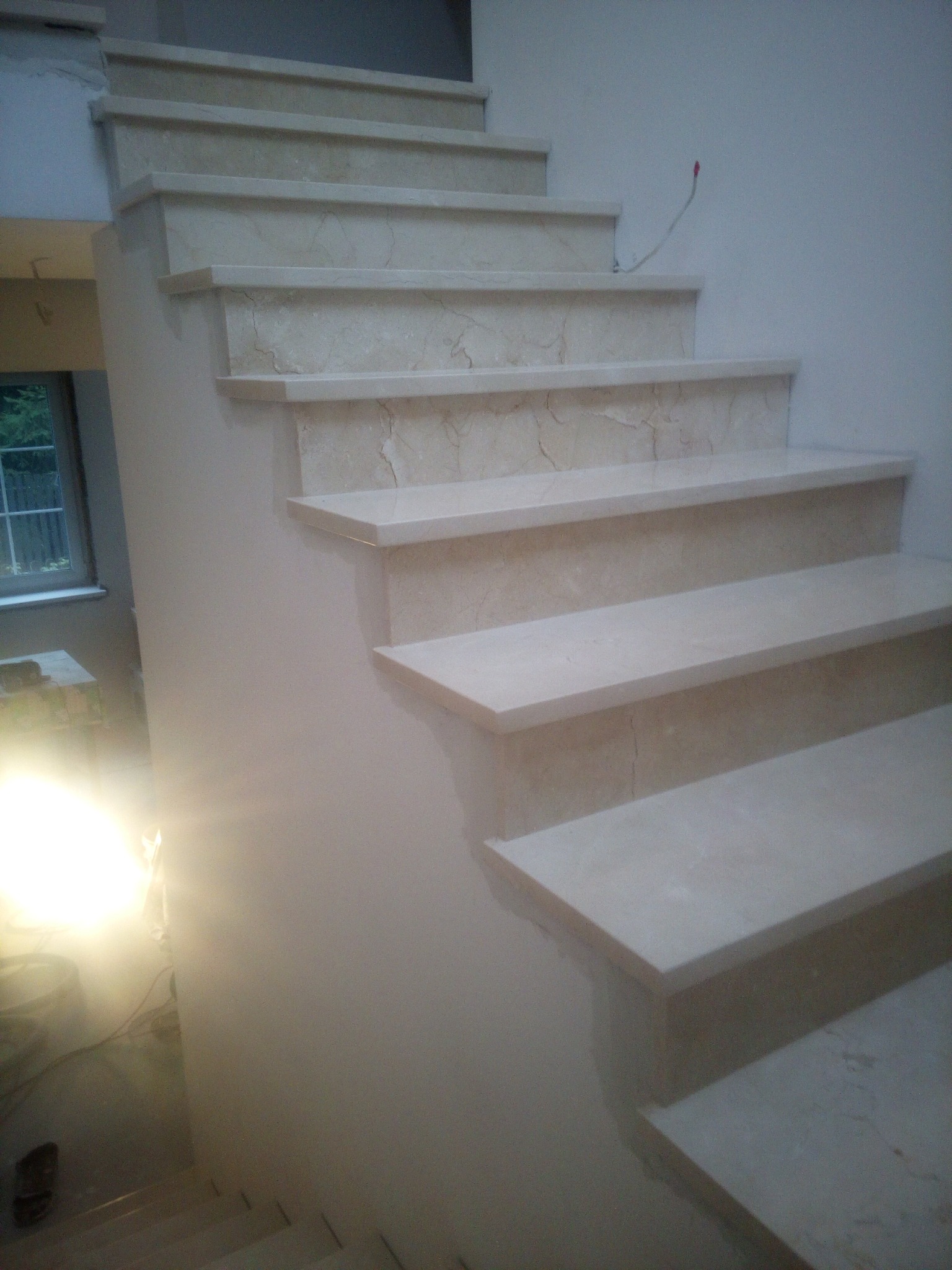 Wewnętrzne schody z jasnego marmuru z widocznymi niedoskonałościami wykończenia i wystającym kablem elektrycznym, w tle okno.