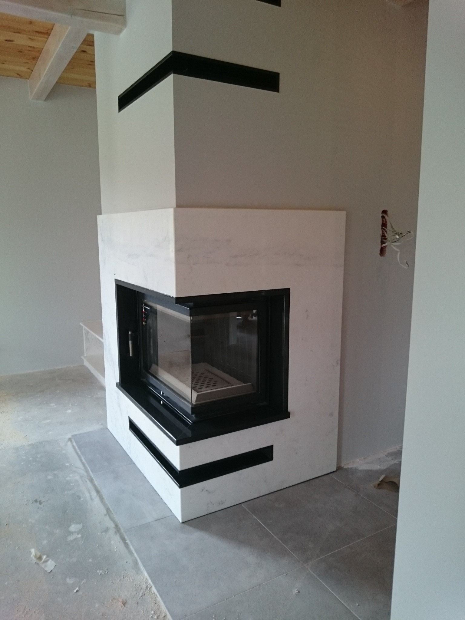 Nowoczesny, narożny kominek obudowany białym kamieniem z czarnymi akcentami, widoczne niedokończone prace wykończeniowe w pomieszczeniu.