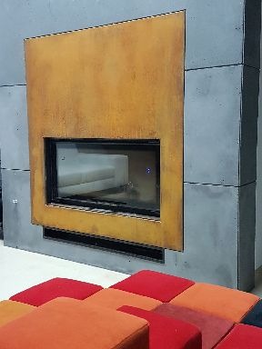 Nowoczesny kominek z obudową z betonu architektonicznego i rdzawego metalu, z prostokątnym wkładem kominkowym i widocznymi kratkami wentylacyjnymi, otoczony kolorowymi pufami.