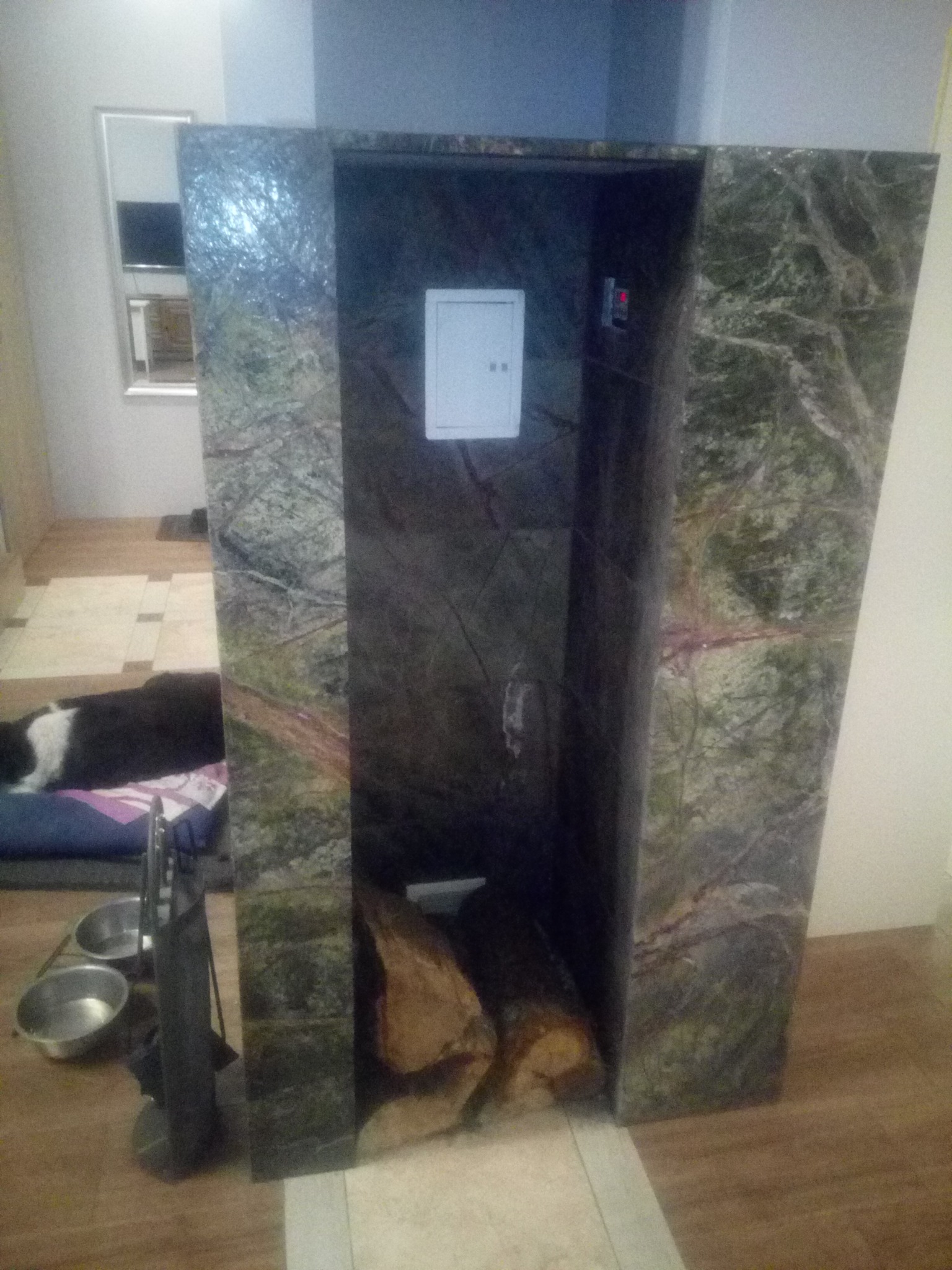 Obudowa kominka z zielonego kamienia z widocznym drewnem wewnątrz, w tle pies leżący na posłaniu i przybory kominkowe