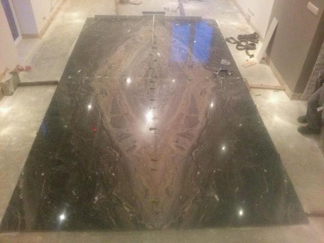 Układanie dużego blatu kamiennego o unikalnym wzorze na podłodze, widoczne narzędzia i elementy montażowe, w tle fragment ściany i okna.