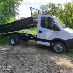 Biały samochód dostawczy z czarną skrzynią ładunkową na błotnistym terenie, widok z boku w słoneczny dzień.