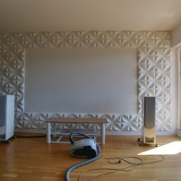 ANDRZEJ BAMBERSKI BYGGESERVICE - Salon z panelami dekoracyjnymi 3D na ścianie, drewnianą podłogą i widocznym odkurzaczem oraz kolumnami głośnikowymi, osoba w tle