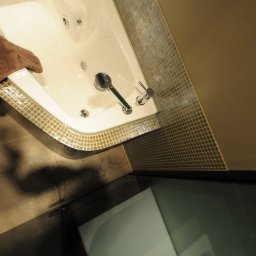 ANDRZEJ BAMBERSKI BYGGESERVICE - Biała wanna narożna z hydromasażem, obłożona mozaiką w odcieniach złota i brązu, z brązowym ręcznikiem na krawędzi. Widoczny element podłogi z butem.