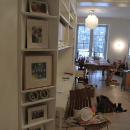 ANDRZEJ BAMBERSKI BYGGESERVICE - Biały regał z półkami wypełnionymi ramkami ze zdjęciami i segregatorami, widok na pokój z dużym oknem, drewnianym stołem i krzesłami, w tle widoczny żyrandol i bałagan