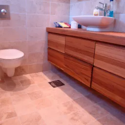 Nowoczesna łazienka z beżowymi płytkami, podwieszaną toaletą i drewnianą szafką pod umywalką w kształcie misy, z widocznym odpływem podłogowym.