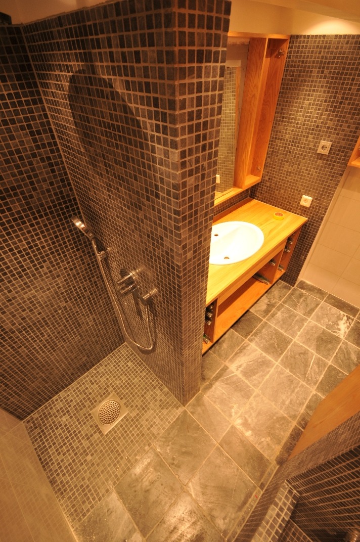 Wykończona łazienka z kabiną prysznicową wyłożoną mozaiką, drewnianą szafką pod umywalką i podłogą z kamiennych płytek.