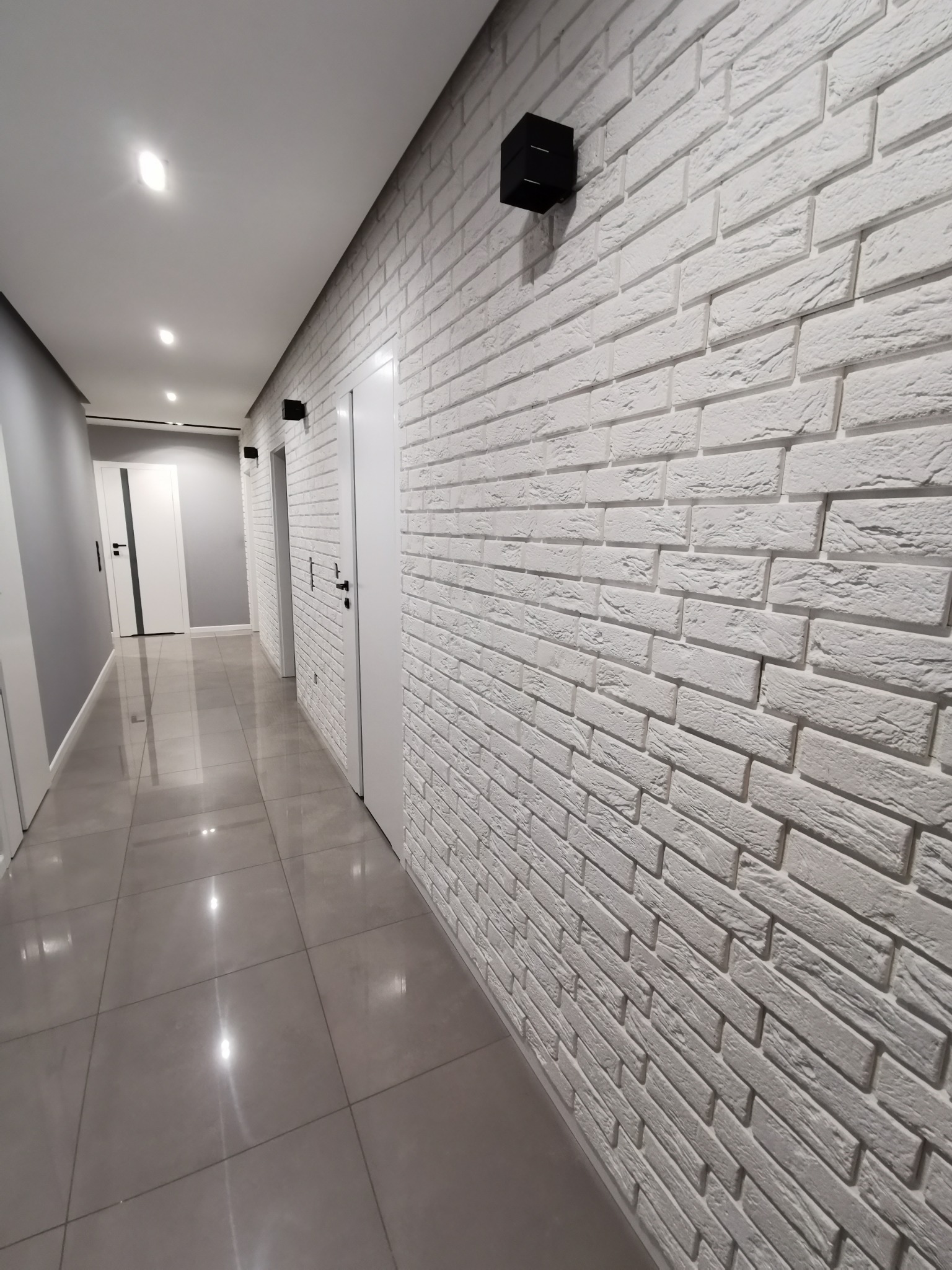 Korytarz z szarą podłogą z płytek i białą ścianą wyłożoną dekoracyjną cegłą, oświetloną czarnymi kinkietami i punktowymi światłami w suficie.
