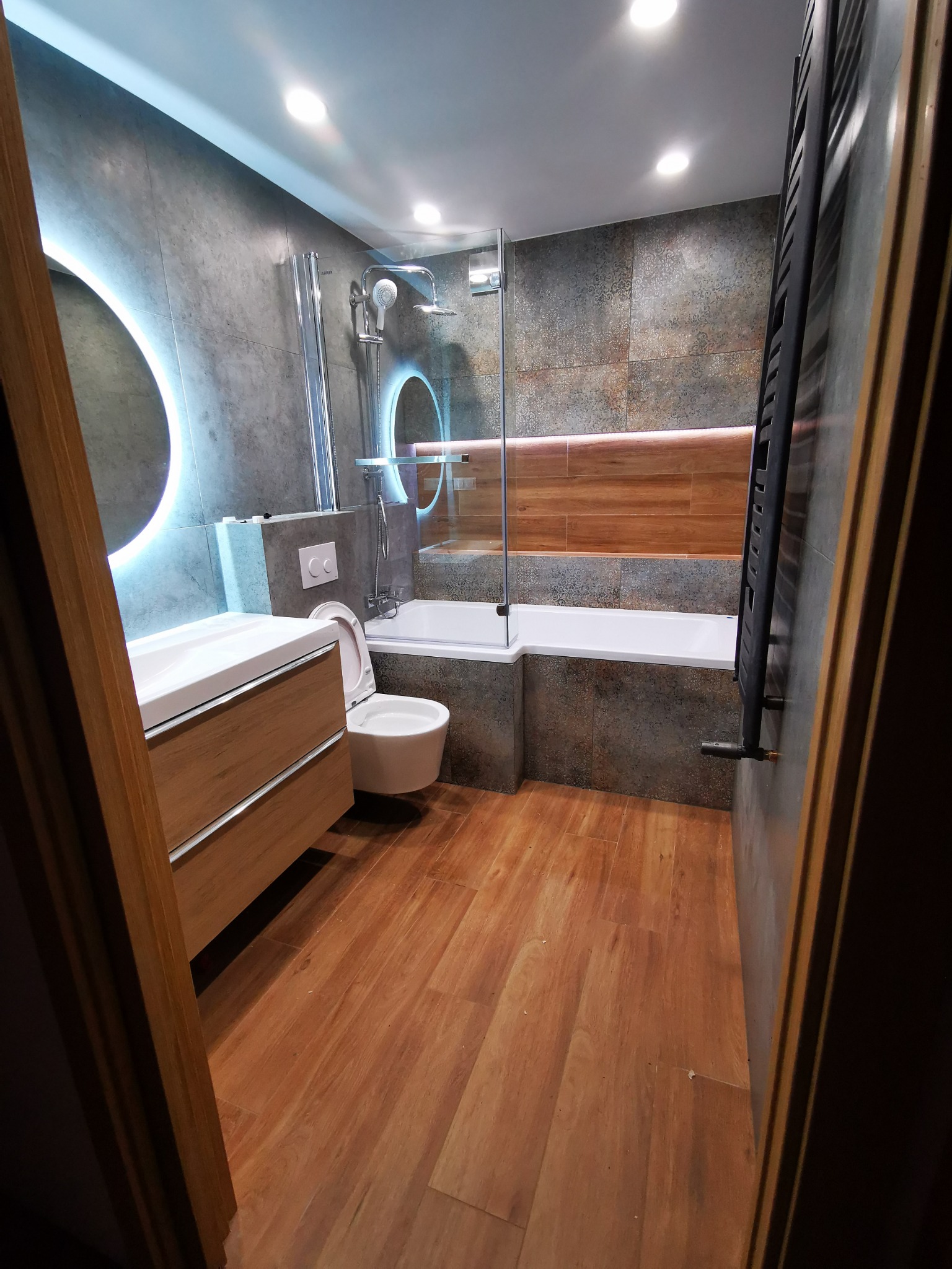 Nowoczesna łazienka z drewnopodobną podłogą i szarymi płytkami, podświetlanym okrągłym lustrem nad umywalką i wanną z parawanem, widok zza futryny drzwi.