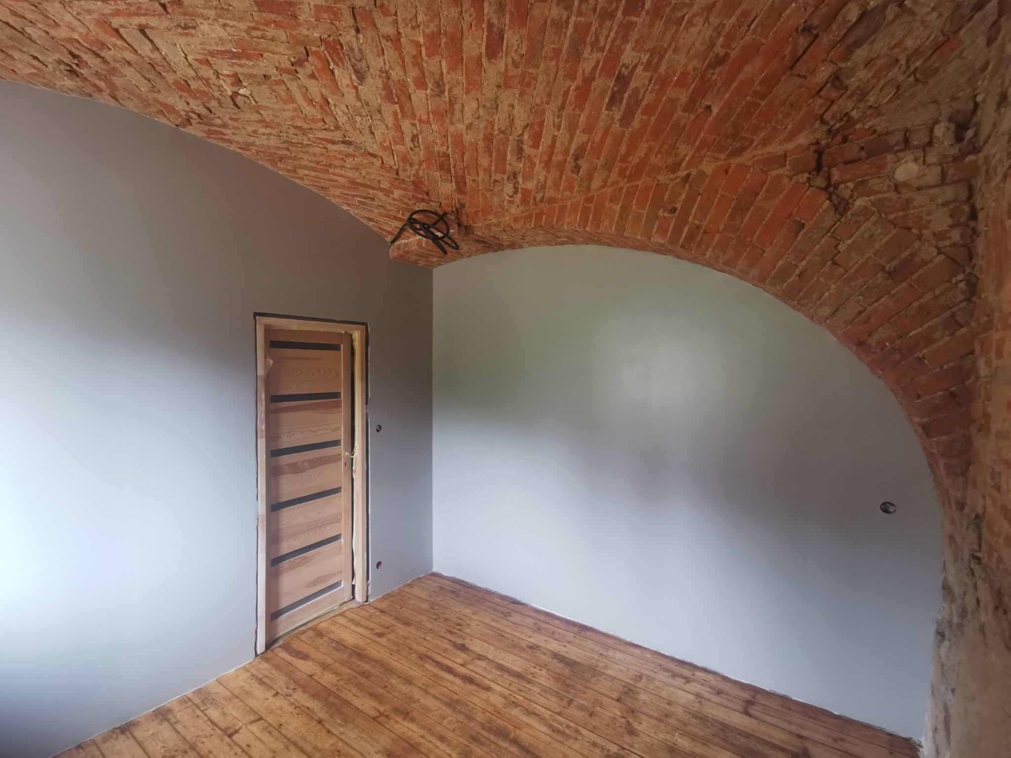 Wnętrze po remoncie: łuk ceglany sufitu, pomalowane szare ściany, drewniana podłoga i nowoczesne drzwi z drewnianymi panelami.