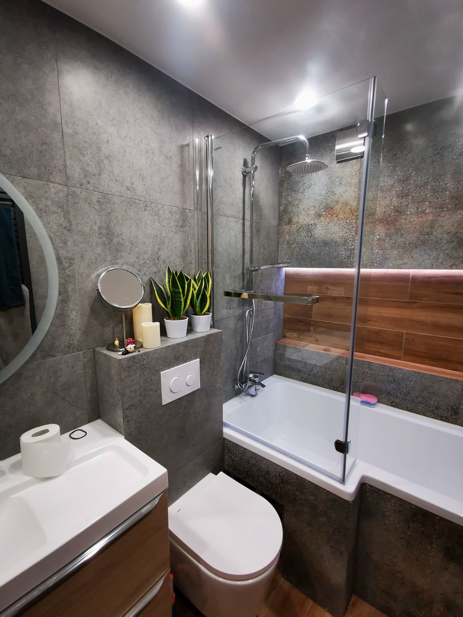 Nowoczesna łazienka z szarymi płytkami imitującymi kamień, białym wyposażeniem, drewnianymi akcentami i roślinami doniczkowymi na podwyższeniu obok toalety.