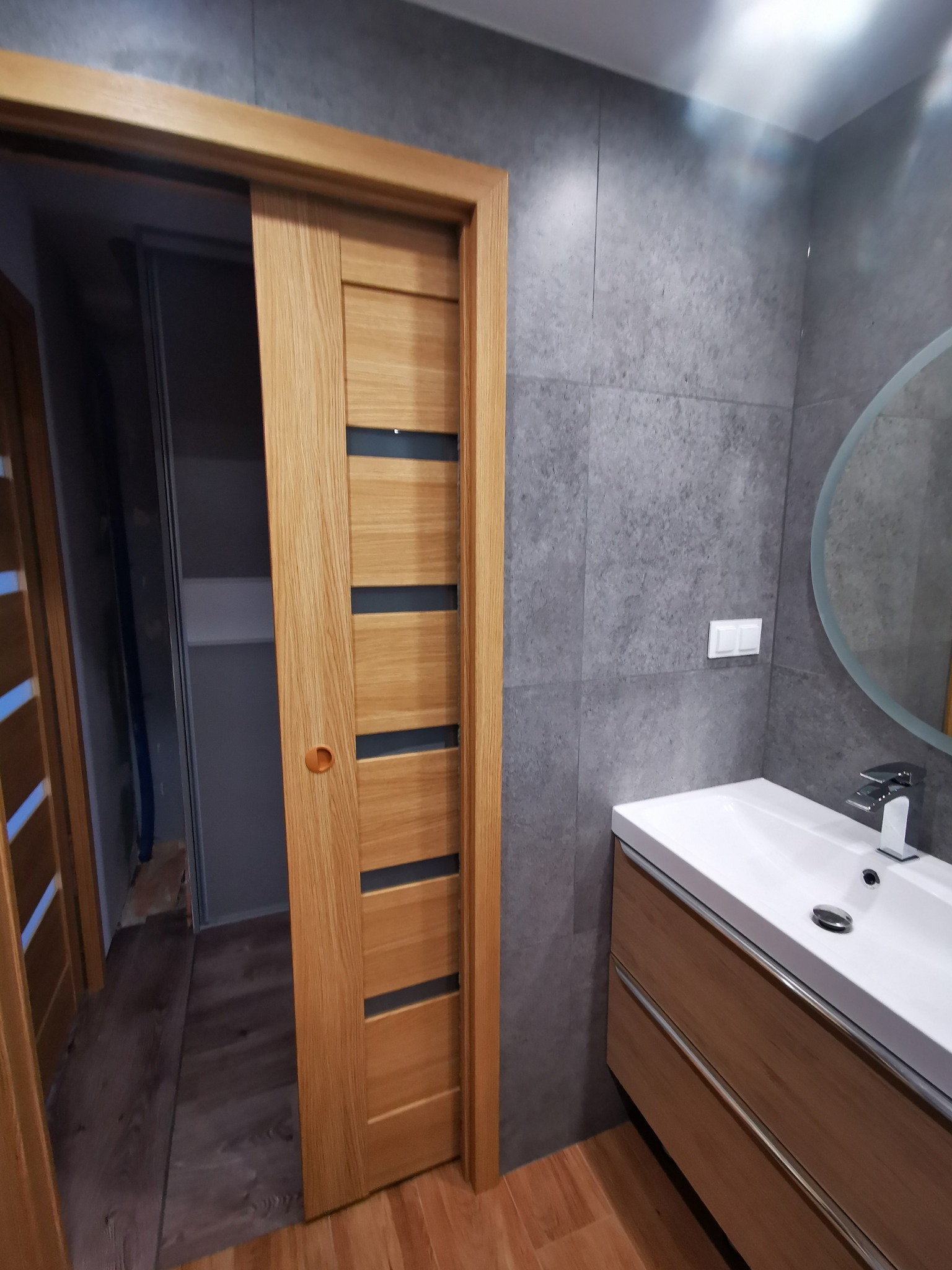 Zamontowane drewniane drzwi przesuwne z czterema poziomymi, ciemnymi wstawkami w łazience z szarą, płytkową ścianą i umywalką z szafką podwieszaną.