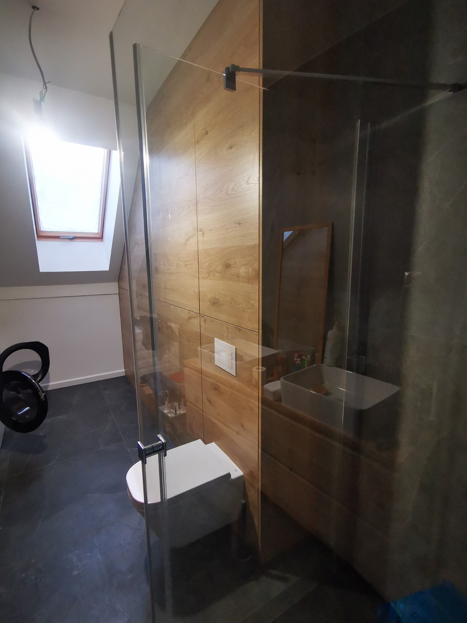 Nowoczesna łazienka na poddaszu z drewnopodobnymi panelami ściennymi, szklaną kabiną prysznicową, umywalką nablatową i oknem dachowym.