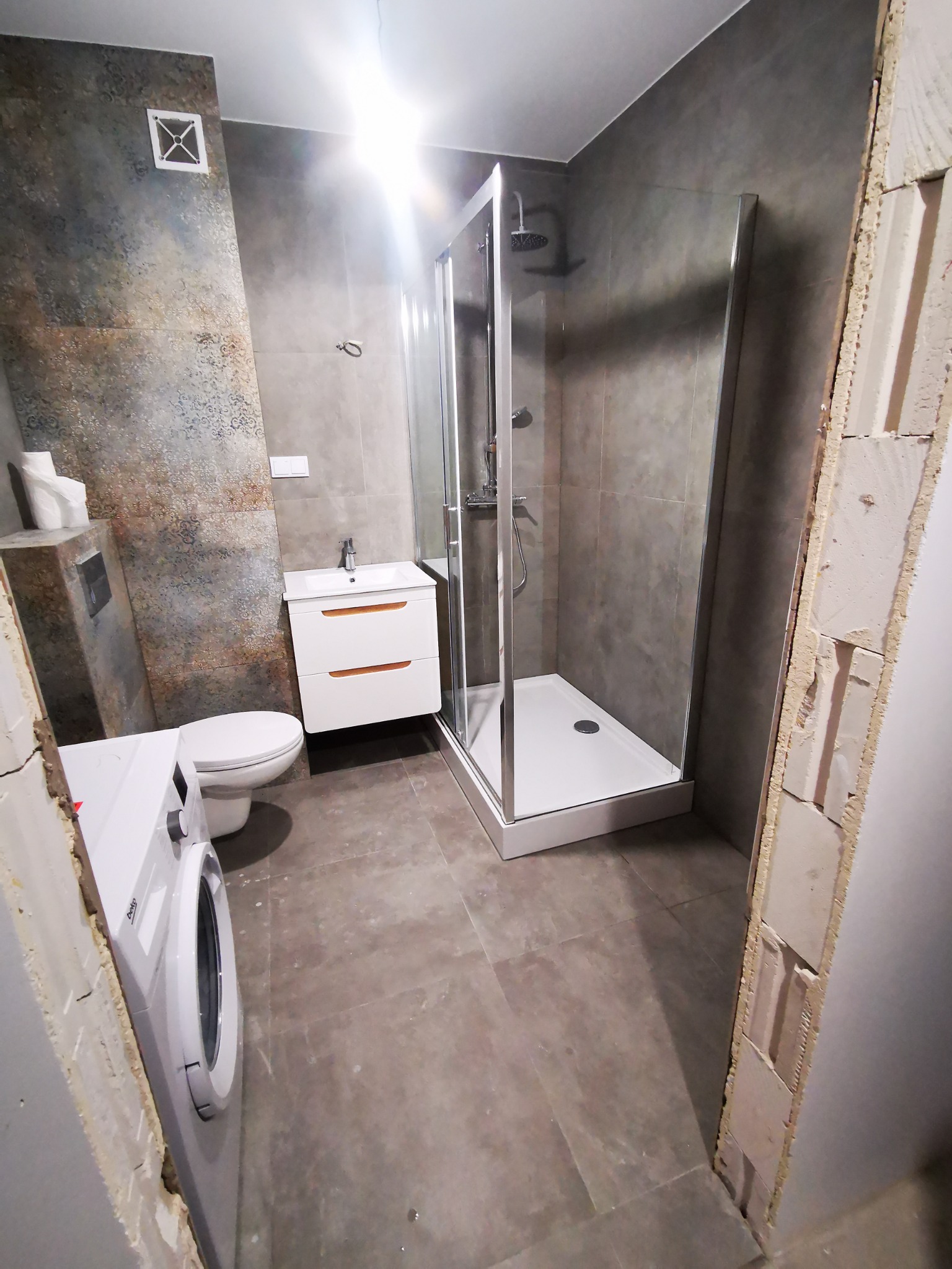 Wykończona łazienka z kabiną prysznicową, umywalką z szafką, toaletą i pralką, ściany wyłożone płytkami w odcieniach szarości i brązu, widoczne surowe wykończenia ścian.