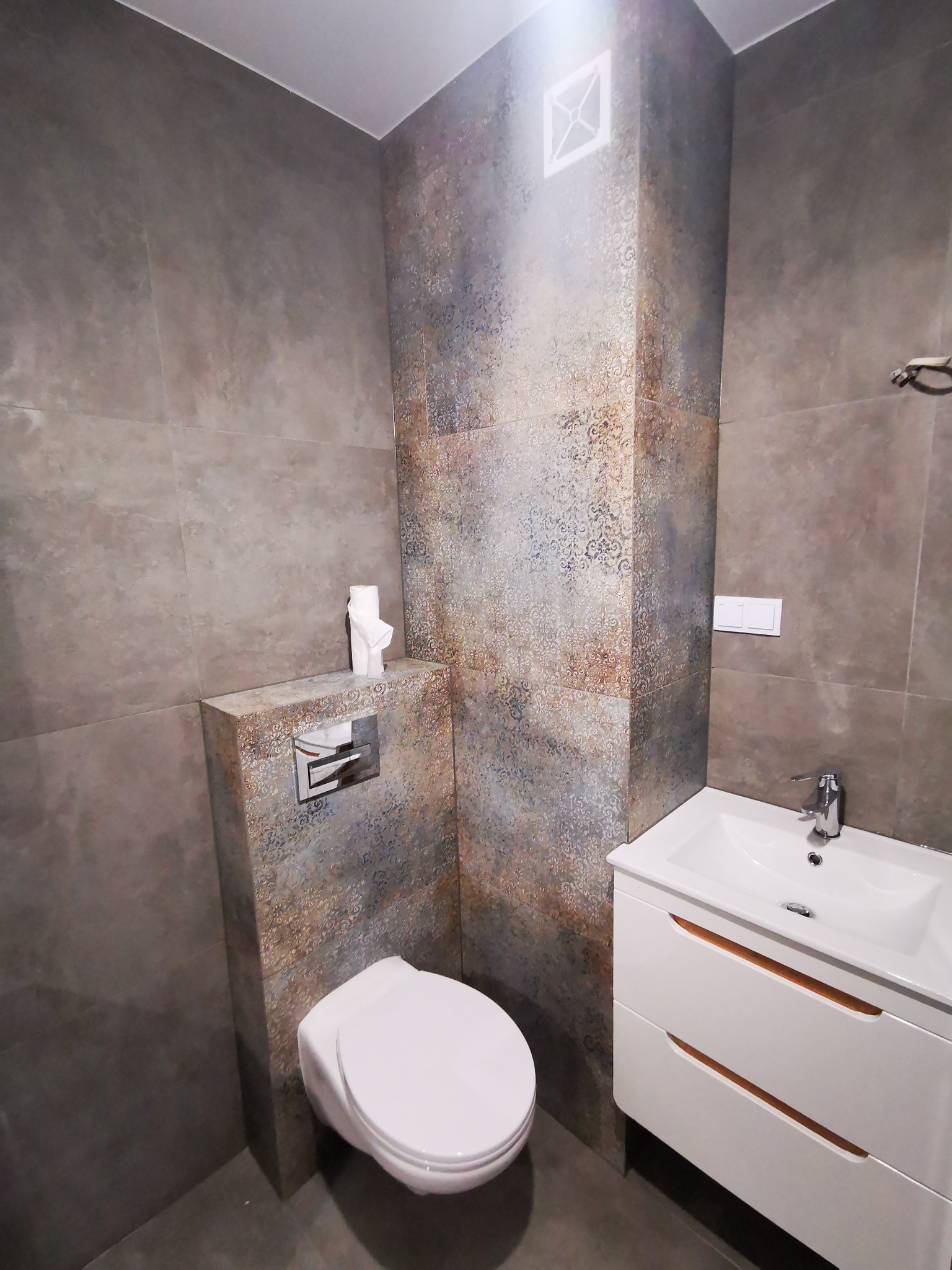 Wykończona łazienka z toaletą podwieszaną, umywalką z szafką w kolorze białym i szaro-brązowymi płytkami z delikatnym wzorem na ścianach i podłodze, widoczne detale armatury i przycisk spłuczki.