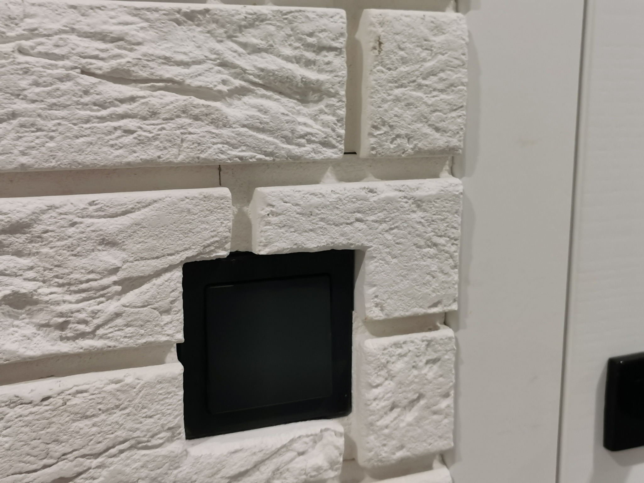 Fragment ściany z białej cegły dekoracyjnej z wkomponowanym czarnym włącznikiem światła, przylegający do białych drzwi z czarną klamką.