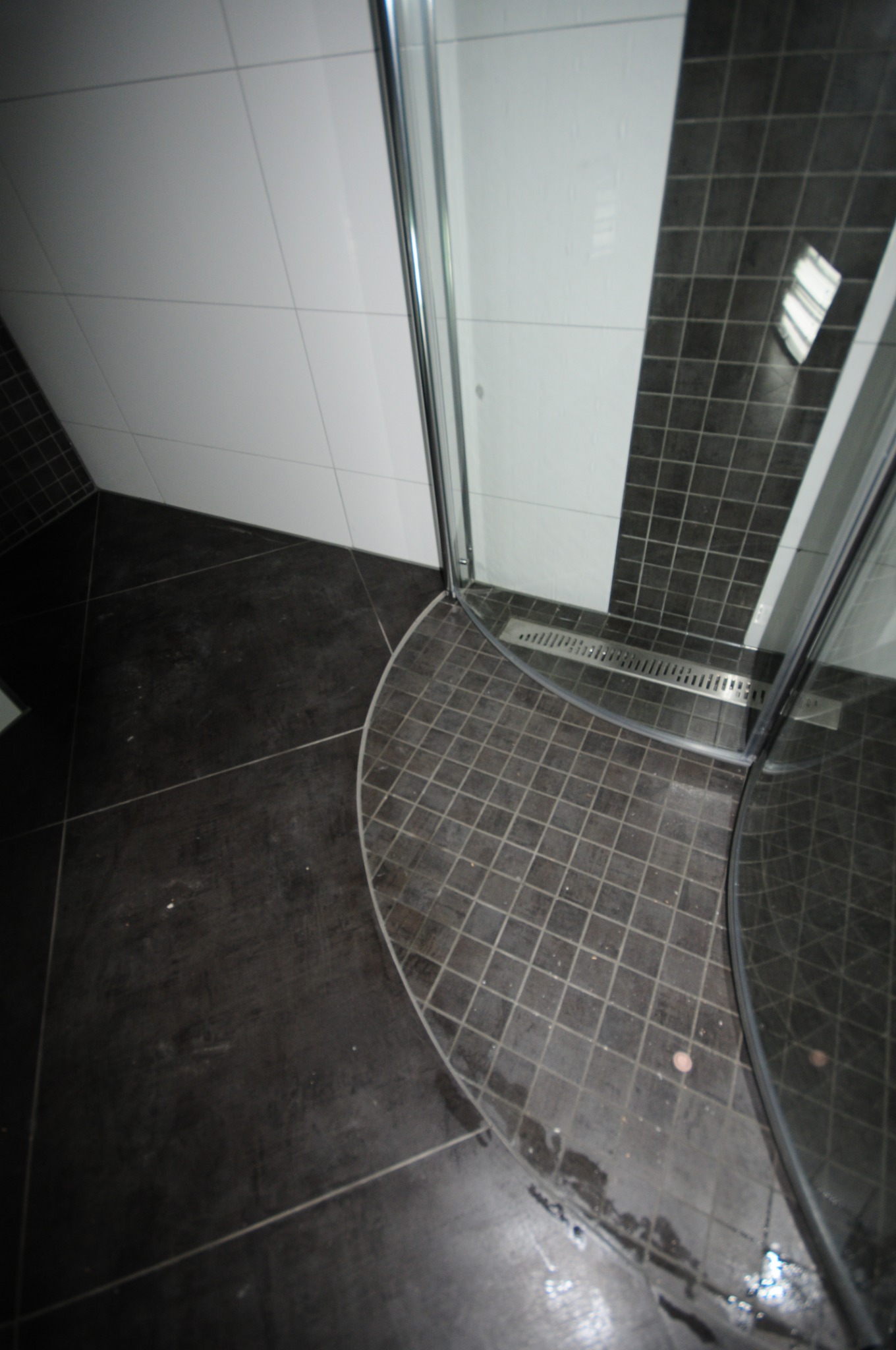 Fragment łazienki z czarnymi płytkami podłogowymi i kabiną prysznicową z odpływem liniowym w rogu, widoczne białe płytki ścienne w górnej części.