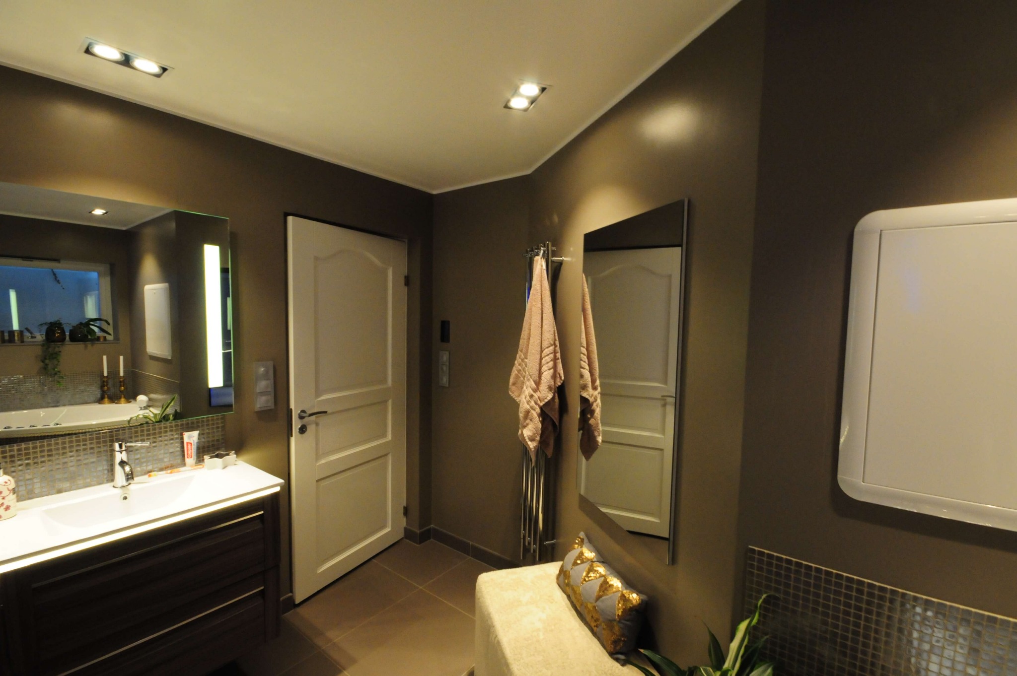 Nowoczesna łazienka z brązowymi ścianami, białymi drzwiami i szafką pod umywalką z ciemnego drewna. Na ścianie wisi lustro, a obok grzejnika ręcznikowego znajduje się pufa z dekoracyjną poduszką.