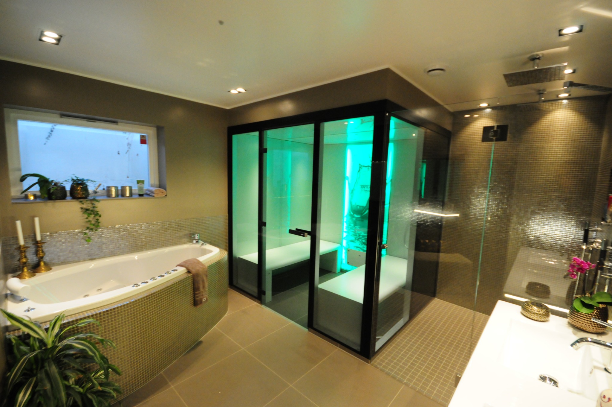 Nowoczesna łazienka z wanną z hydromasażem, kabiną prysznicową typu walk-in i szklaną sauną z zielonym oświetleniem, wykończona mozaiką w odcieniach brązu i beżu.
