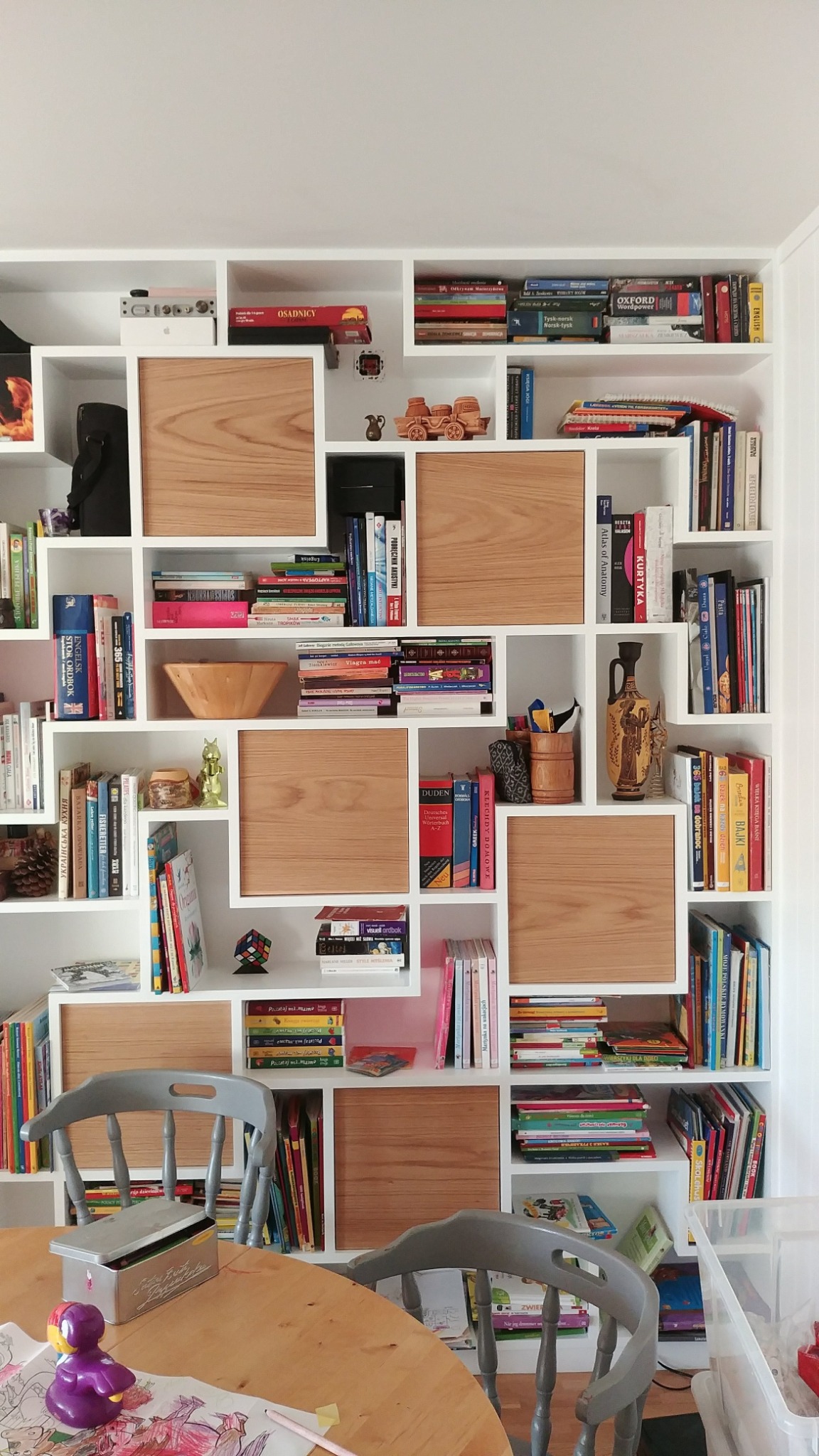 Biały regał z drewnianymi wstawkami, wypełniony książkami, ozdobami i zabawkami, widoczny zza drewnianego stołu z szarymi krzesłami, na którym leżą kredki i rysunki.