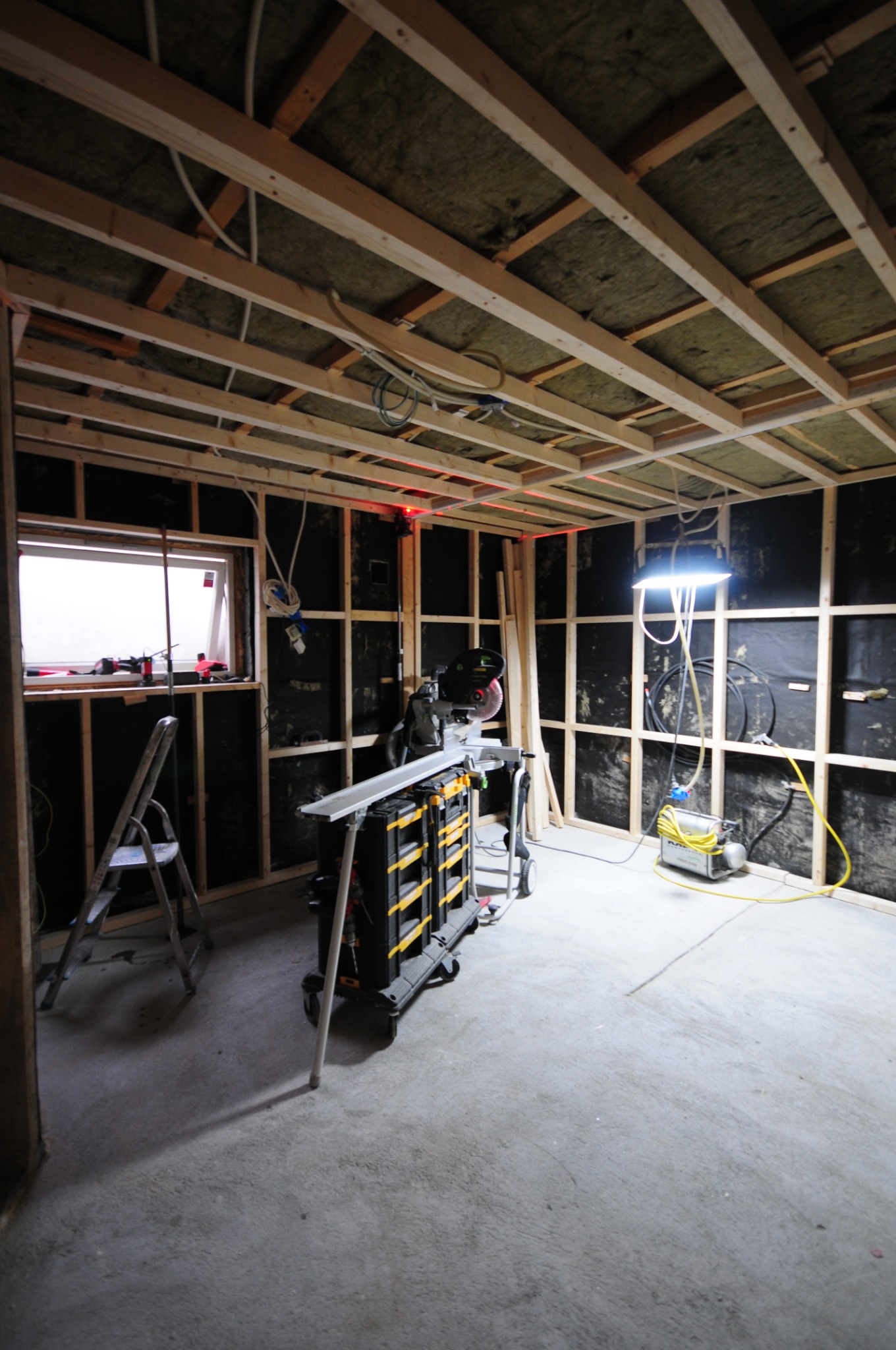 Wnętrze w trakcie prac: widoczne drewniane ramy ścian, izolacja stropu, narzędzia budowlane, ukośnica na wózku, drabina i oświetlenie robocze.