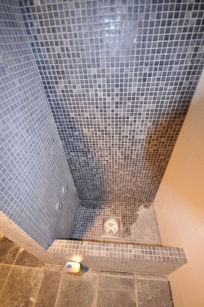 Wnętrze kabiny prysznicowej wyłożonej drobną, szarą mozaiką, z widocznym odpływem liniowym i dwoma okrągłymi punktami na ścianie, prawdopodobnie wlotami wody.