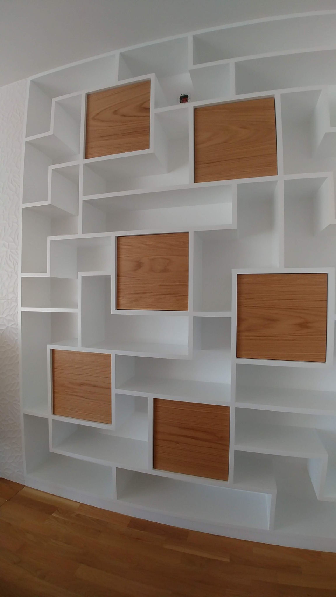 Biały, asymetryczny regał z drewnianymi wstawkami, wbudowany w ścianę z dekoracyjną fakturą, widoczny fragment drewnianej podłogi.