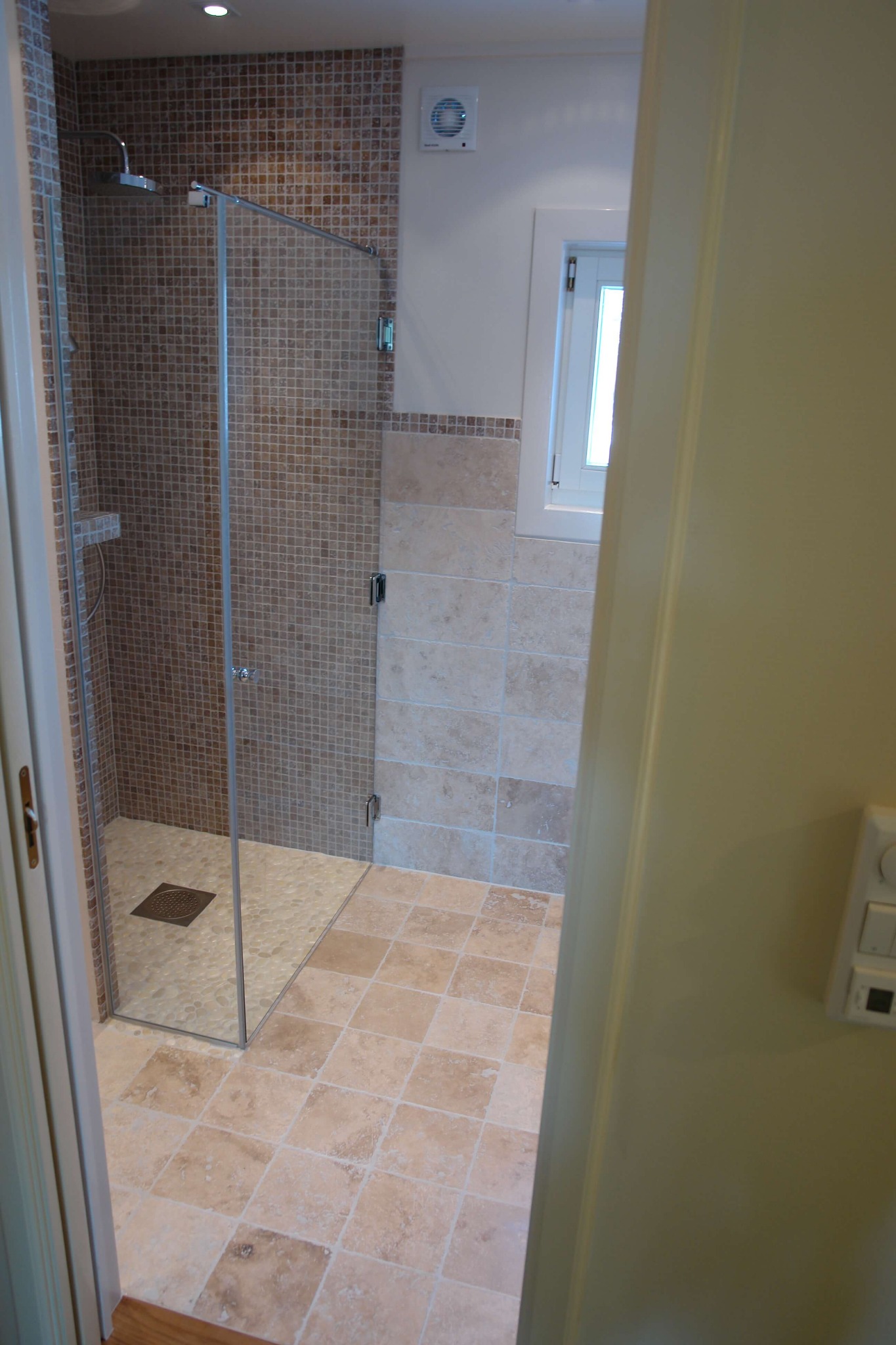 Wykończona łazienka z kabiną prysznicową typu walk-in, wyłożona beżowymi płytkami mozaikowymi i kwadratowymi, widoczny fragment okna i wentylator.