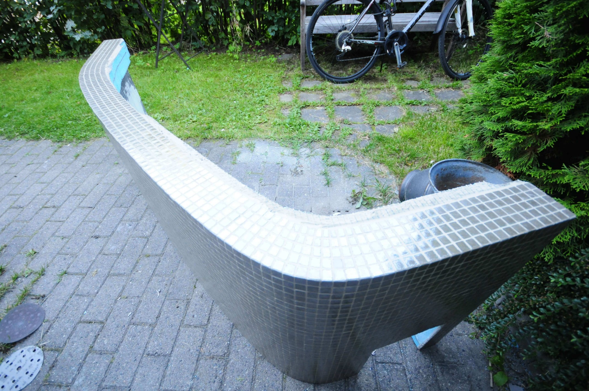 Niedokończona, zakrzywiona konstrukcja obłożona mozaiką ceramiczną, z widocznymi fragmentami niebieskiego podkładu, leżąca na brukowanej powierzchni w ogrodzie, z rowerem i zielenią w tle.