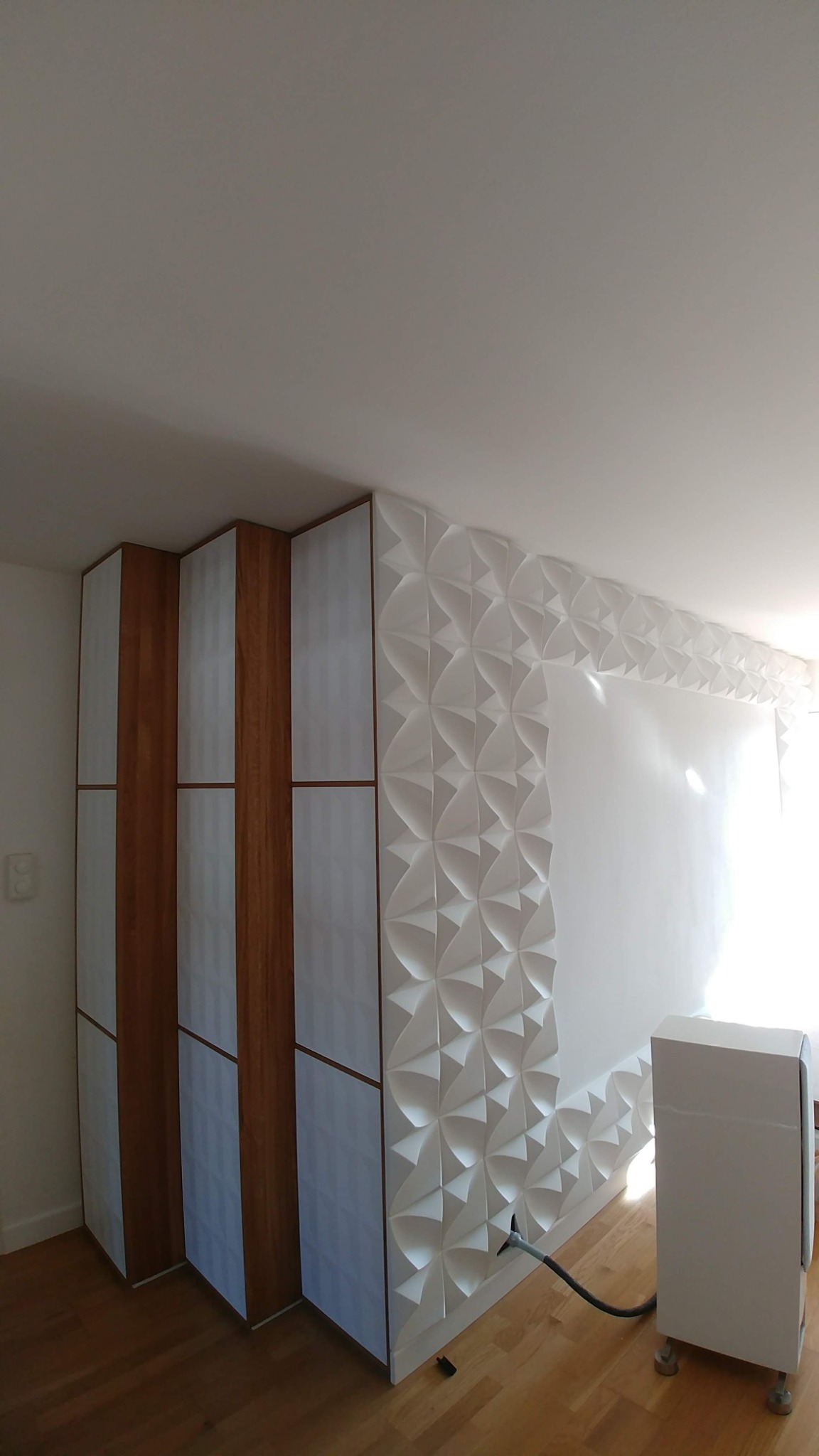 Nowoczesne wnętrze z zabudową meblową z drewnianymi akcentami i dekoracyjną ścianą z białych paneli 3D, oświetlone naturalnym światłem.