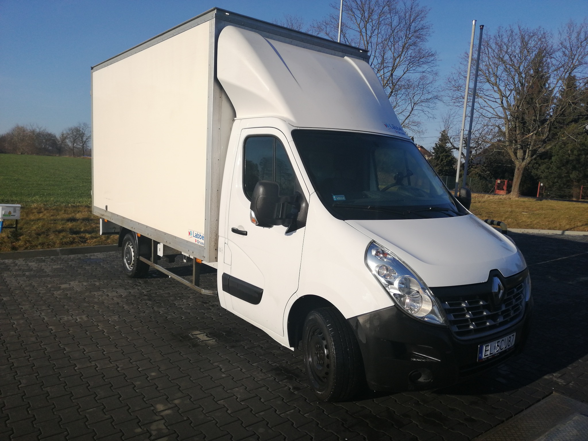 Biały samochód dostawczy Renault Master z zabudową kontenerową, zaparkowany na kostce brukowej, widoczny bok i przód pojazdu, w tle pole i drzewa bez liści pod błękitnym niebem.