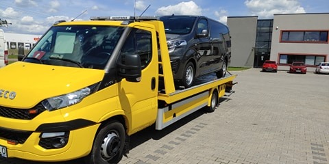 Żółta laweta Iveco z szarym busem na platformie, widoczna w tle budynek biurowy i zaparkowane samochody.