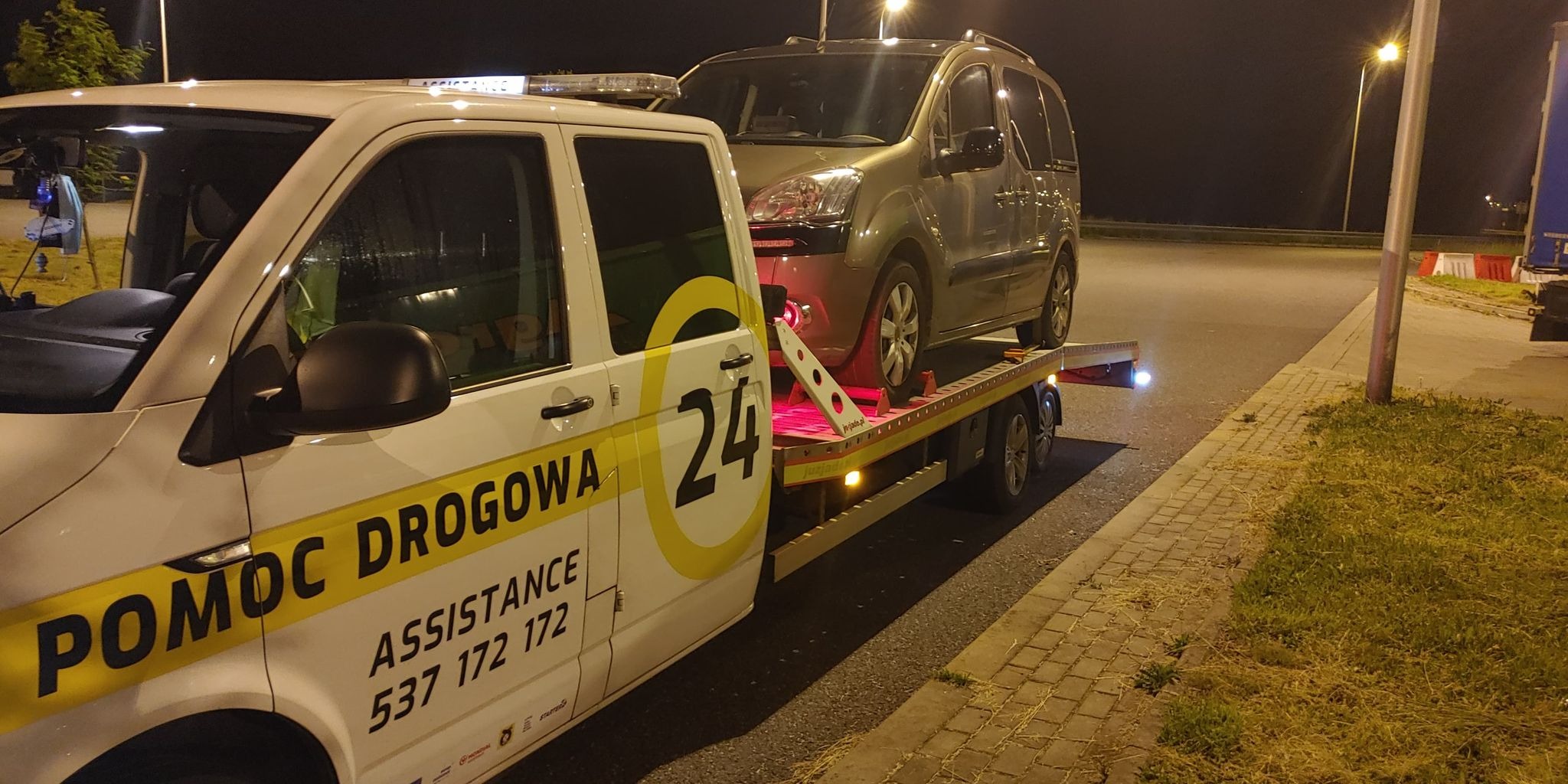 Samochód osobowy koloru khaki przewożony na lawecie pomocy drogowej z widocznym numerem '24' i napisem 'POMOC DROGOWA ASSISTANCE 537 172 172' podczas nocnej akcji na oświetlonej ulicy.
