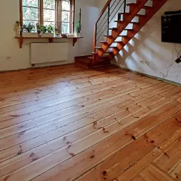 Wycyklinowana podłoga z desek w jasnym odcieniu drewna, widoczne schody drewniane z czarną balustradą, okno z roślinami w tle, ściany białe, gniazdka elektryczne widoczne na ścianie.