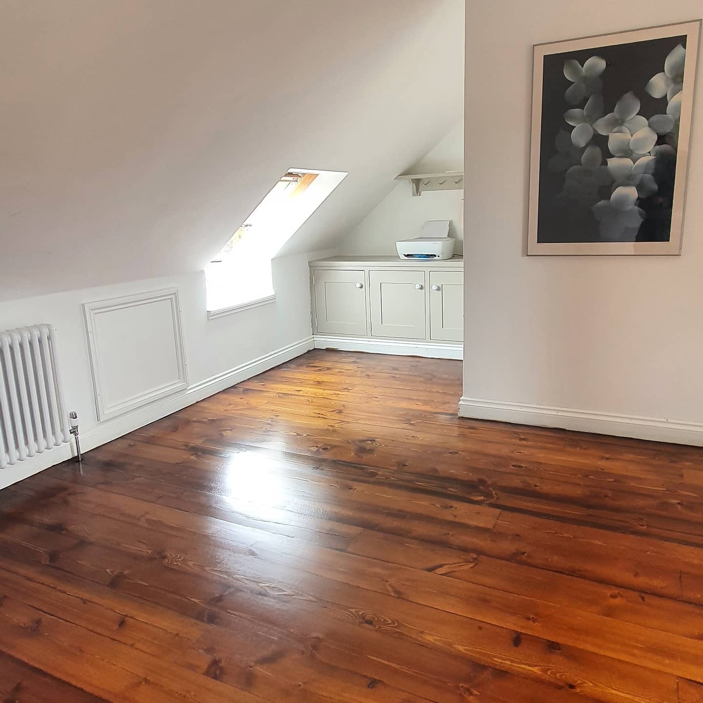 Poddasze z odnowioną, ciemnobrązową podłogą z desek, widoczne okno dachowe, białe ściany, grzejnik żeberkowy i szafka z drukarką w tle oraz obraz na ścianie.