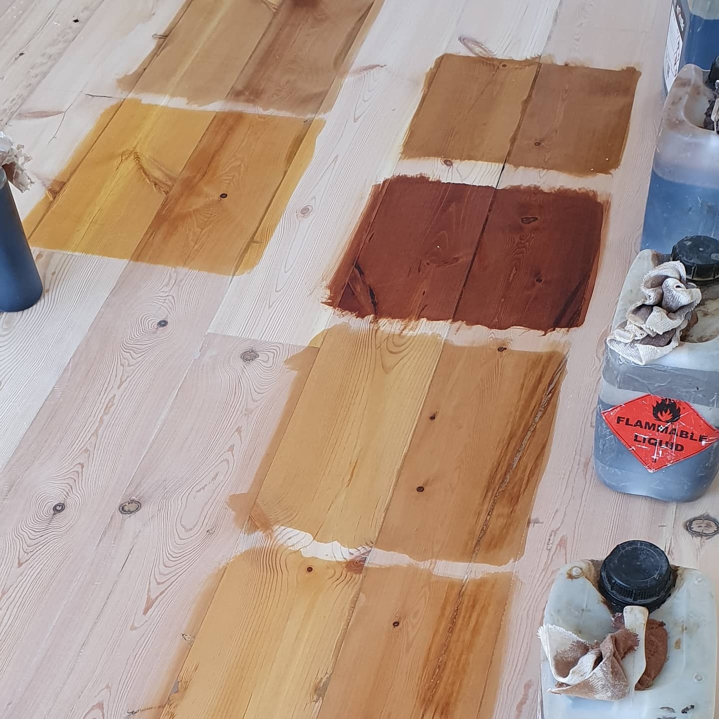 Testowanie różnych odcieni bejcy na surowej drewnianej podłodze, widoczne pojemniki z bejcą i szmatki