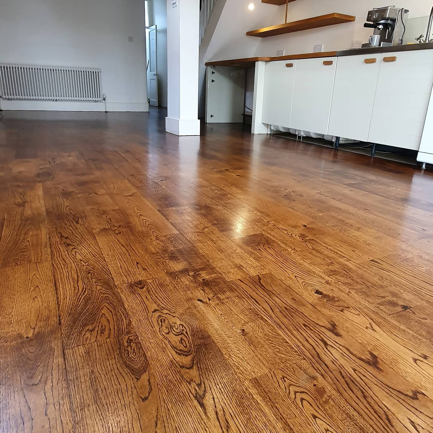 Błyszcząca, brązowa podłoga z desek w jasnym wnętrzu z białą zabudową kuchenną w tle, widoczny grzejnik i kolumna podtrzymująca schody.