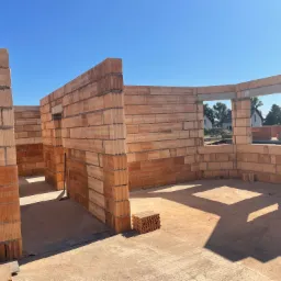 Wnętrze domu w stanie surowym otwartym, wzniesione z czerwonej cegły ceramicznej, widoczne otwory okienne i drzwiowe, konstrukcja w trakcie realizacji.