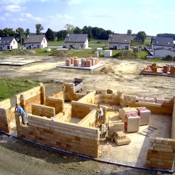 TOMASZ WITEK WT BUDOWNICTWO - Budowa domu jednorodzinnego w trakcie wznoszenia ścian z bloczków ceramicznych, widoczne fundamenty pod kolejne budynki, teren budowy z pojazdami i materiałami budowlanymi, perspektywa z lotu ptaka.