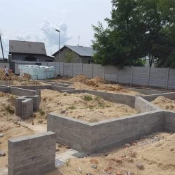 TOMASZ WITEK WT BUDOWNICTWO - Wykopane i częściowo wymurowane fundamenty pod budynek mieszkalny, widoczne wystające pręty zbrojeniowe i hałdy piachu, w tle ogrodzenie z prefabrykatów betonowych oraz budynki mieszkalne.