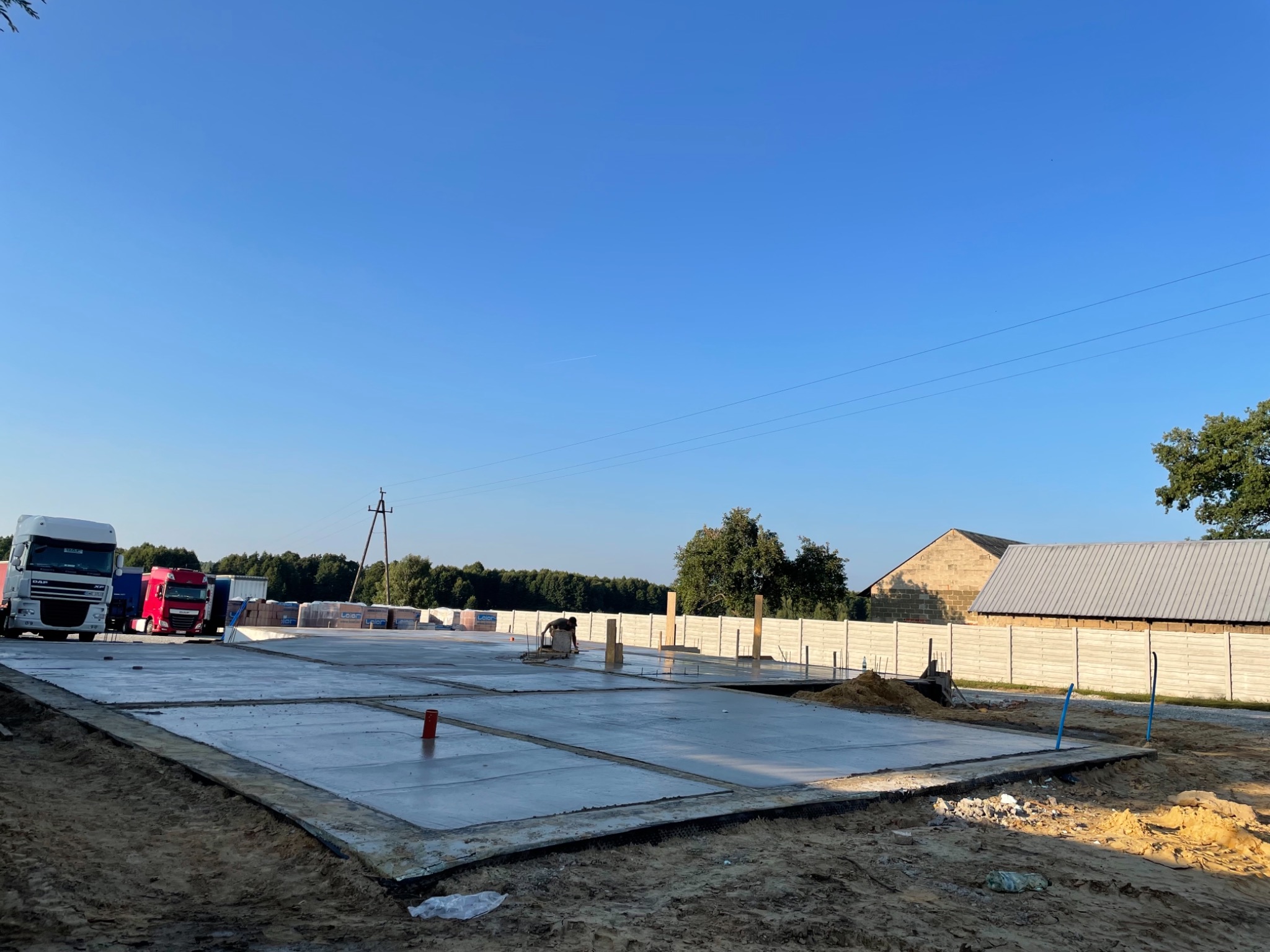 Wylany fundament z zaznaczonymi sekcjami, na placu budowy widoczne ciężarówki dostawcze i materiały budowlane, w tle drzewa i budynek gospodarczy.