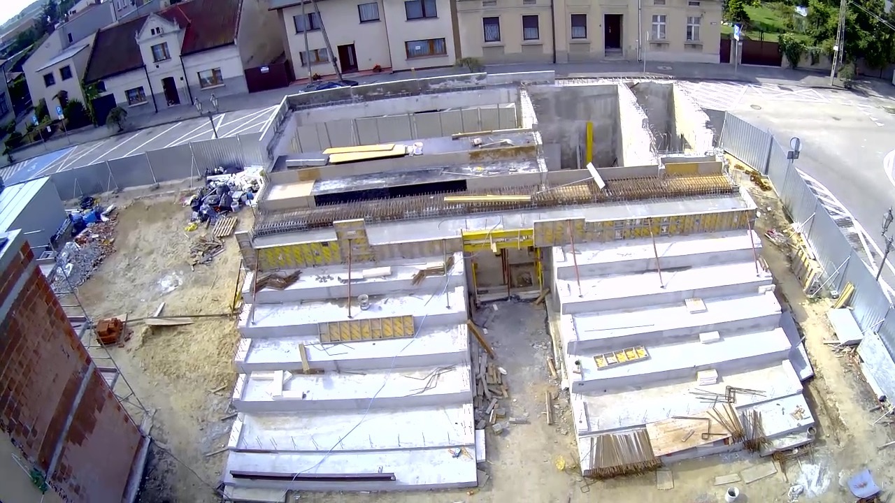 Widok z góry na plac budowy z wykopami pod przyszłe budynki, widoczne przygotowane szalunki i zbrojenia, częściowo zalane betonem, w tle zabudowa miejska.