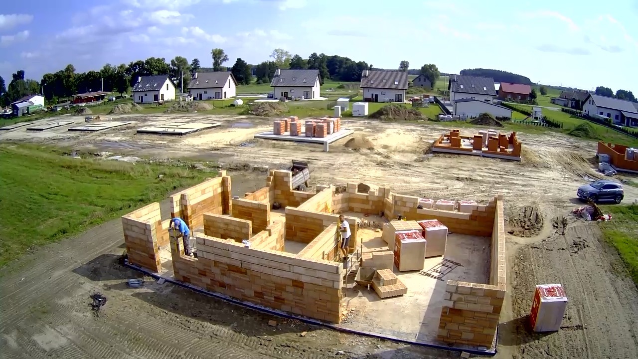 Budowa domu jednorodzinnego w trakcie wznoszenia ścian z bloczków ceramicznych, widoczne fundamenty pod kolejne budynki, teren budowy z pojazdami i materiałami budowlanymi, perspektywa z lotu ptaka.