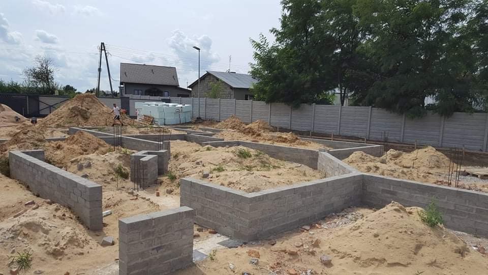 Wykopane i częściowo wymurowane fundamenty pod budynek mieszkalny, widoczne wystające pręty zbrojeniowe i hałdy piachu, w tle ogrodzenie z prefabrykatów betonowych oraz budynki mieszkalne.