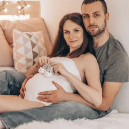 Przyszła mama i tata siedzą na łóżku, trzymając małe, urocze buciki dziecięce na tle brzuszka, za nimi dekoracyjne lampki.