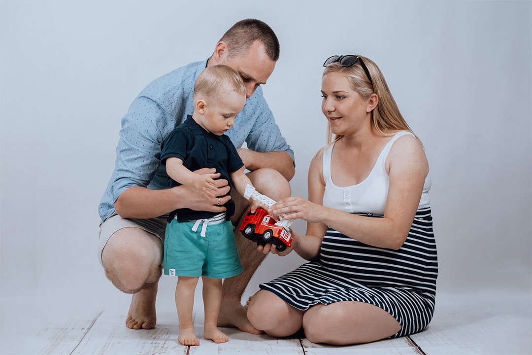 Przyszła mama w sukience w paski siedzi na podłodze i bawi się z synkiem, trzymając w rękach czerwoną zabawkę w kształcie wozu strażackiego, obok niej stoi ojciec w niebieskiej koszuli.