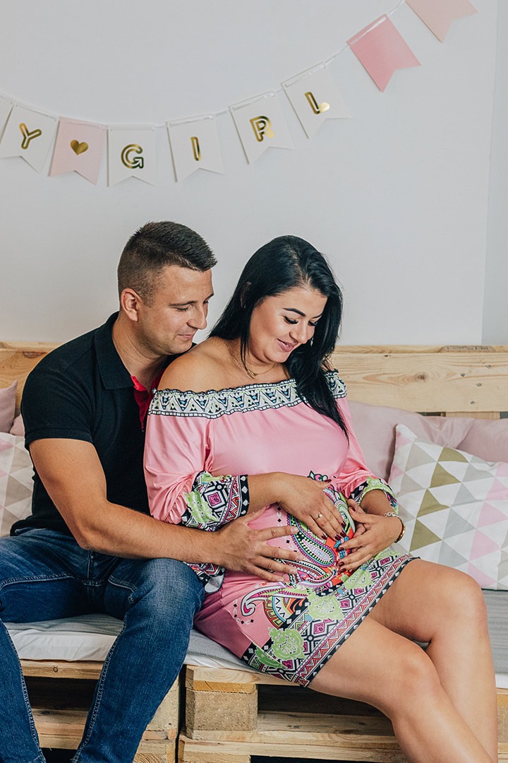 Przyszli rodzice siedzą na łóżku z palet, trzymając ręce na brzuchu kobiety w wzorzystej, różowej sukience, nad nimi wisi transparent z napisem 'Y <3 GIRL'.
